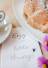 Tasse Kaffee und ein Stück Kuchen mit Puderzucker auf einem Teller, dekoriert mit Blumen, dazu eine Karte mit der Aufschrift ‚Enjoy the little things
