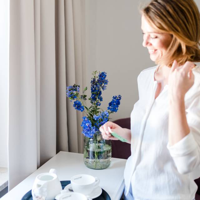 Frau steht lächelnd am Tisch mit Blumen und Getränk
